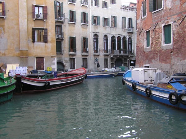 Boats parked along a canal in Venice, Italy.  The boats are open, and look like they are used for work, about 20 feet long and painted in bright colors, blue, red, and white.  They are floating in slightly murky greenish water.  Above the boats are the walls of buildings surrounding the canal.