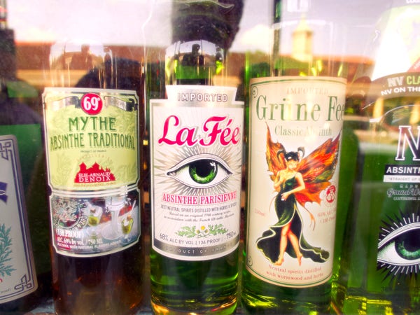 Landscape orientation, color, photo close up to the liquor store front window. Five bottles of absinthe are lined up, but only three are fully in frame. the bottles are emerald green and from left to right there is a bottle of Mythe Absinthe Traditional, then a bottle of La Fee absinthe with an emerald eye on the label, and then a bottle of Grüne that features of pixie like figure with an emerald dress ponytails, and what appears to be orange wings. Some of the window reflections hint at the skyline behind me. But not much is distinguishable. The photo has a slight dream like quality.