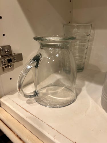 a glass pitcher on a shelf, with a hinge on the left suggesting this is a cabinet. There's also a glass that matches the pitcher in the background. It seems like a simple and clean design, suitable for serving beverages