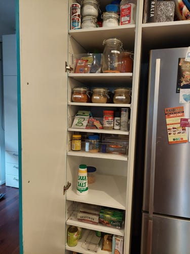 Tall cupboard open showing shelves with things neatly stored on them. One shelf has 3 glass jars with neat, printed labels on the jars. The nearly empty middle shelf has a tall container of salt and one small plastic container on it.