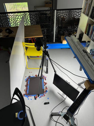 A piece of black foamcore taped to the floor with blue tape. A tripod straddles it with a camera on top plugged into a monitor and remote trigger. A large LED ceiling light rests to the right propped up on saw horses and 4x4s. A large white reflector sits opposite precariously leaning on ikea folding chairs