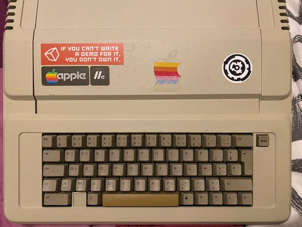 An Apple IIe with a sticker saying “If you can’t make a demo for it you don’t own it.” and a Revision sticker.