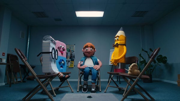 A still image of a short film made entirely with puppets, featuring a young character called Geoff in a group therapy session with other puppet characters based on his favorite everyday tools, including a notepad, a pencil, an eraser and a laptop.
