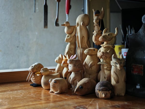 Photo of a group of woodcarved creatures : a walrus, a duck with a butter knife, a grumpy grog, a standing cat, a standing fox, a elephant eared shrew, a hedgehog, a three eyed gargoyle, a kobold, a standing t rex, a dragon with a round duck on its head, a humanoid octopus, a monster with a red robin on its head and a round rabbit in its left hand, and a weird creature with a plump rabbit as its head