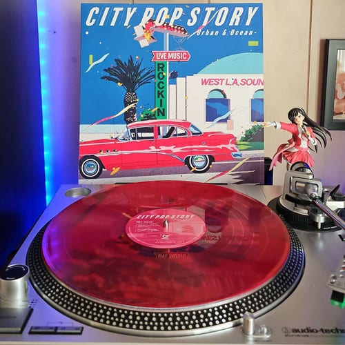 A transparent red vinyl record sits on a turntable. Behind the turntable, a vinyl album outer sleeve is displayed. The front cover shows artwork of a classic car in front of a building and signs. 