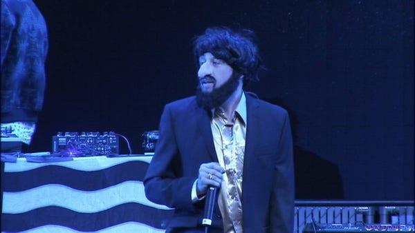 Mackelmore onstage with mic dressed as a Jew caricature. Dark suit, black wig, fake beard, big prosthetic nose.