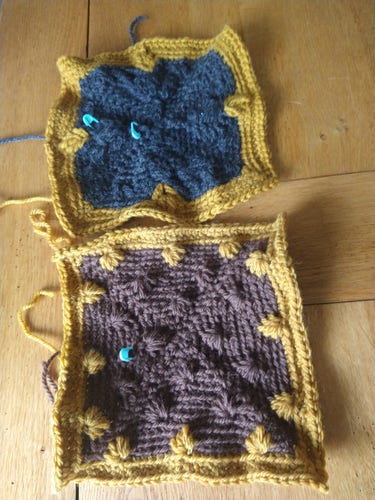 Two crochet squares on a table at the top is bluey green with a yellow border, below is a brown centre with yellow border in a bobble pattern 