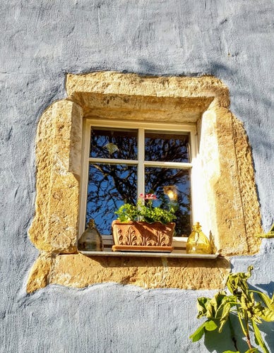 Ein Fenster Umrandung sind Sandsteinblöcke. Die Häuserwand ist blau. Auf dem Fenstersims steht ein pflanzkasten aus Ton mit grünen 
 Pflanzen. In der Fensterscheibe ist das Spiegelbild eines  Baumes zu sehen.