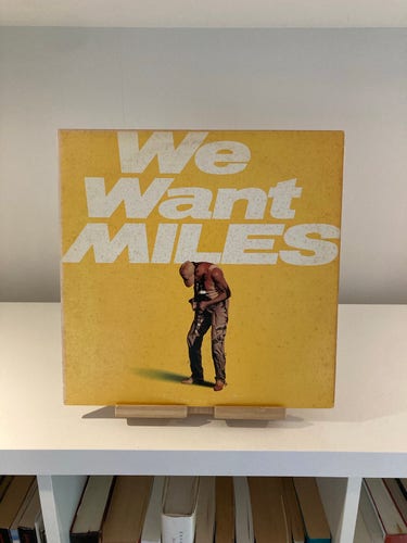 An album cover titled "We Want Miles" displayed on a shelf, with a picture of Miles Davis holding a trumpet and large text over a yellow background. Below the album, a row of books on a shelf is visible.