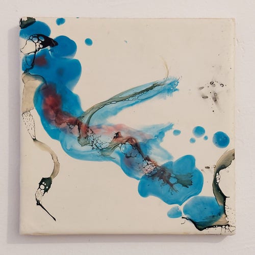 a piece of square wood used as a canvas for an encaustic abstract painting made on top of it. This painting has a painted white background that is noticeable enough because it gives space for the colors to breathe. There's a trail of organic blue going from the top left to the bottom right of the canvas, and inside of that blue there's hints of blue and on top of it some black paint that was burned and gives different thin-shaped patterns in specific parts of the painting.
