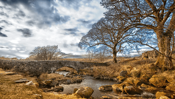 Pastoral landscape image of a stream flowing through an abandoned slate works with a limestone bridge int he middle distance. There are sycamore trees lining the stream and the landscape gives way to a partly clouded bright winter sky.