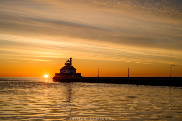 The sun rises over Lake Superior and the Duluth lighthouse. Everything is yellow and orange in color.