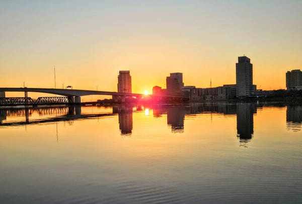 Glowing orange and yellow sunrise over a vast river with silhouettes of bridges and a city skyline on the opposite shoreline with the bright sun rising between the silhouettes, all reflecting their image upon the calm river below.