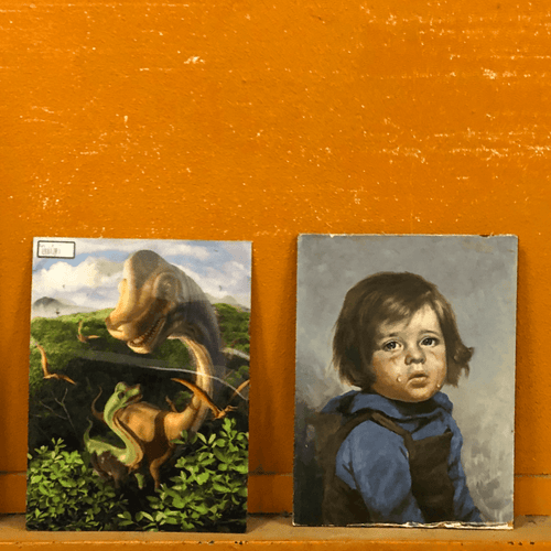Two small unframed images near the bottom of the frame aganst a dark orange wall. The image on the left is of a large and small dinosaur in the scene of a jungle/forest. The image on the right is of a small boy with brown hair, blue shirt and dark overalls, looking sadly outward at the viewer.