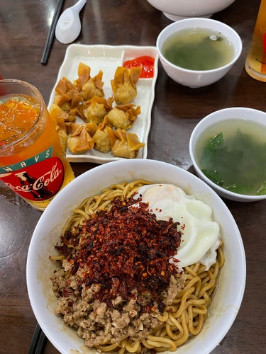 A bowl of noodles lightly stirred in dark sauce and topped with minced meat, an egg and lots of dried chili flakes. Around it are two bowls of vegetable soup, a glass of Aiyu jelly, and some deep-fried dumplings.