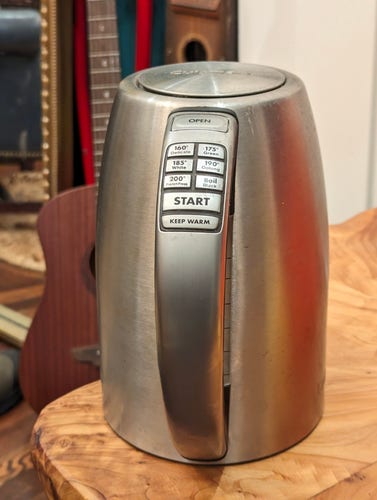 A KitchenAid variable temperature kettle (it has multiple buttons allowing the desired temperature to be chosen, making it easier to steep different types of tea) sitting on a wooden table. In the background is a ukulele next to a mirror.