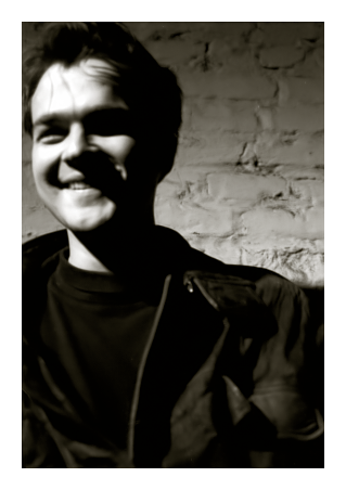 black and white, low-angle close-up snapshot lit by a single bulb. a mid-20s man with dark hair, tee and jacket, smiling broadly. he's standing in front of a brick wall painted white, leaning away and looking left, his face slightly out of focus. 