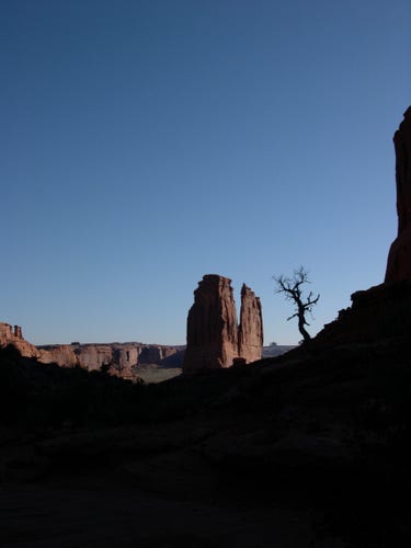 As darkness curves across the bottom 40% of the view and into the right margin, beyond the darkness a rock formation stands in front of a deep valley and a cerulean sky, and next to the rock formation stands the silhouette of a blasted tree.