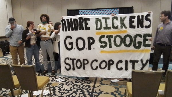 Protesters lock arms at the front of a conference roon, holding a banner reading Andre Dickens GOP Stooge. Stop Cop City 