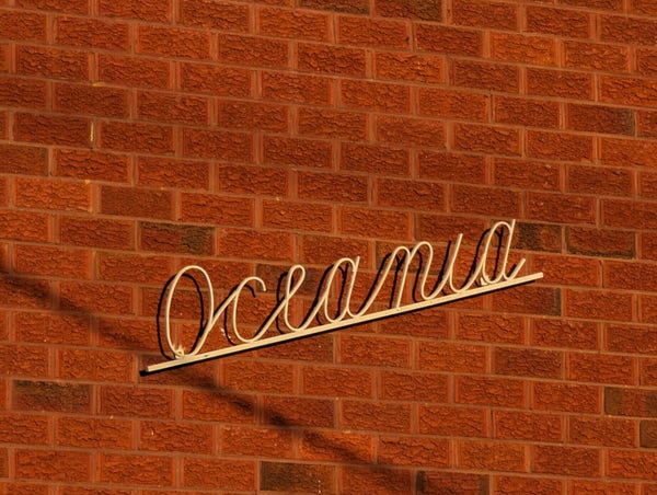Red brick wall in late evening sun shining against a golden metal cursive sign saying 'oceania' as the name of the building in a gorgeous piece of art deco slash modernism style