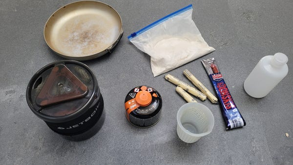 A jetboil, lightweight titanium pan, a bag of dry damper mix, three cheese sticks, a packaged pepperoni, olive oil bottle and small measuring cup, all on a grey stone bench top. Oregano, garlic powder and sachet of tomato paste not shown
