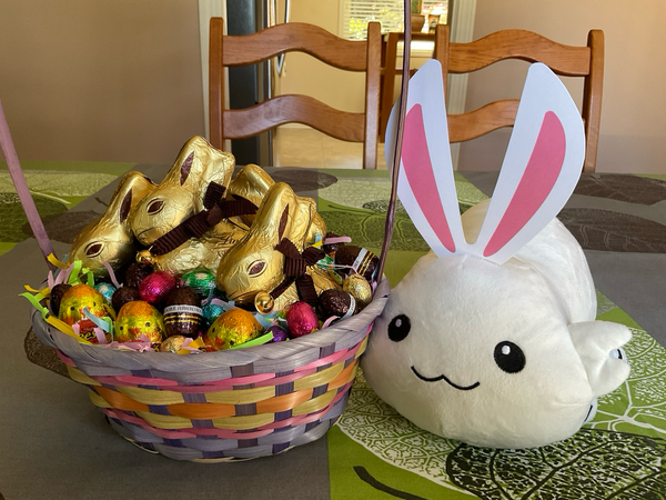 Plush moopsy wearing paper rabbit ears, next to an Easter basket filled with Lindt chocolate bunnies, Reese peanut butter eggs, Ferrero Rocher chocolate eggs, and mini chocolate eggs.