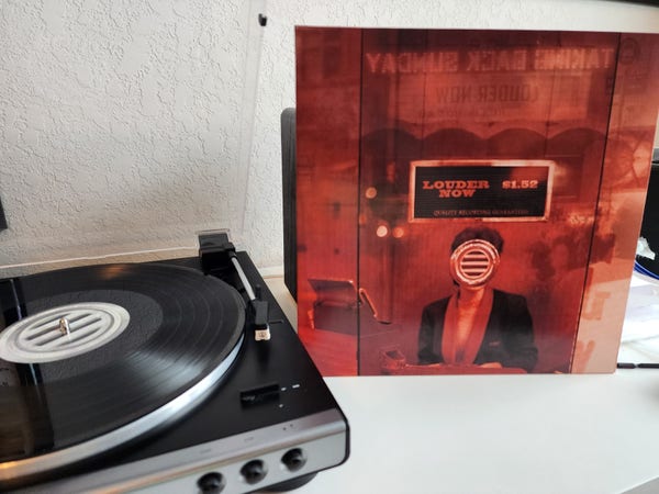 Taking Back Sunday - Louder Now album cover next to a record player. Cover is red, an image of a woman in a ticket booth, her face obscured by the talk box. Vinyl is black.