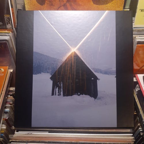 Lp box cover features the photograph entitled "Haus mit Rakuten (House with Rockets)" by Roman Signer. It's a picture of a small, weathered wooden sided structure in a snowy, mountainous setting in what appears to be the early dusk. Two long beams of light appear to be shooting off of the roof in intersecting lines of light, heading up to the sky.