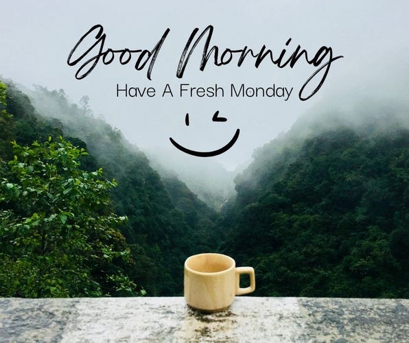 The image features a serene, misty mountain landscape viewed from a high vantage point. In the foreground, there's a ledge with a simple beige coffee mug placed on it. The background shows lush green mountains partially shrouded in fog, creating a tranquil and refreshing atmosphere. Overlaid on the image is the text "Good Morning, Have A Fresh Monday" in an elegant, cursive script. Below the text, there's a smiley face drawn with a simple line for a mouth and two dots for eyes, adding a cheerful touch to the message.