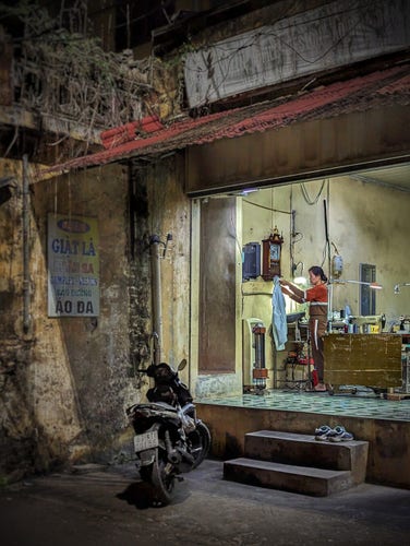 An evening photo of the entrance to a one-room tailor and laundry in a village of Hưng Yên province. We're standing in the street looking into the slightly elevated first-floor business. The space is recessed a bit, with a sign on a neighbor's exposed side wall that advertises laundry services (and the various types of clothing they can clean). A single motorbike is parked out front by a pair of concrete steps leading up to the workshop. There, we see a woman standing with a white shirt in her outstretched hands. She is in front of a workbench with a sewing machine atop it. An antique clock is mounted on a worn,  mold-splotched wall of what was once yellow paint in a high ceilinged room.