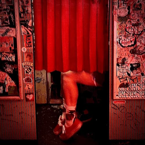 Outside of a photo booth with two people inside taking photos. The one closest to the opening is wearing shorts and sneakers. The curtain is red and the room is also illuminated in red.