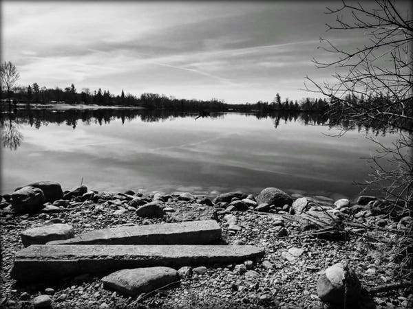 A black and white image of a rocky shoreline beside a small lake. Large rocks and trees can be seen as well as the reflection of clouds in the calm water.