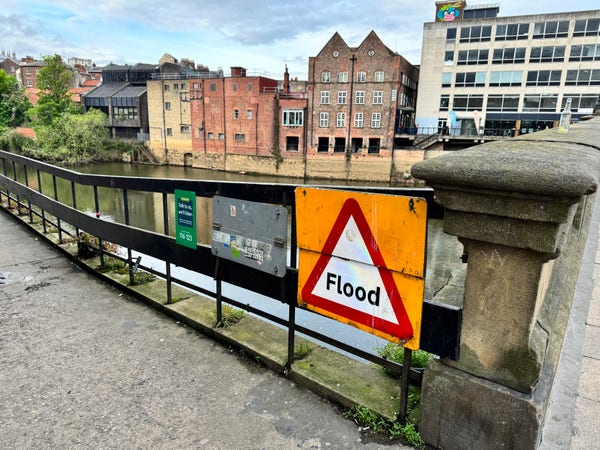 A large warning sign mounted on the railing next to the River Ouse just says “Flood”