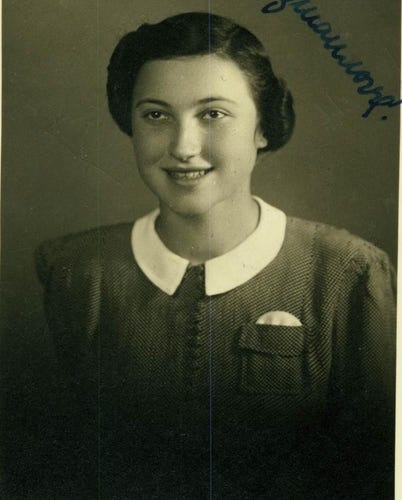 Portrait photograph for documents of a young woman. Her torso and head are visible. She has medium length hair curled into waves and pinned back. On the right side of her head - a parting. She smiles and exposes her upper teeth. She is wearing a fine cardigan blouse with a semi-circular collar. On the right side on her chest a pocket from which a white round handkerchief protrudes.