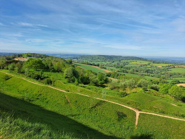 View looking over the ramparts of Painswick Beacon hillfort. Multiple lines of banks and ditches below, the green grass cut through to the limestone beneath along the tops where people have walked. Green and yellow patchwork fields below, with distant hills fading to blue.