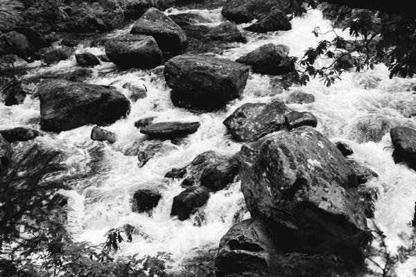 A black and white photograph of stones on a fast flowing river. The stones are grey and black and the water a frothing and pristine white.