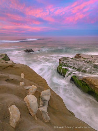 Vertical seascape photo showing pink pre-sunrise clouds over the ocean and coastal rock formations in the foreground. On the left are strange concretions that are embedded into softer sedimentary rock, on the right a small, momentary "tidefall" flows from rocks into the surf.