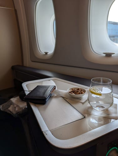 A first class seat on a BA A380, a glass of water and some nuts are on the table, and there are two windows.