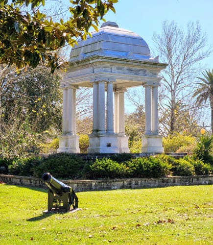 A small black cannon sits on lush green grass in a well manicured park next to a large white stone gazebo.