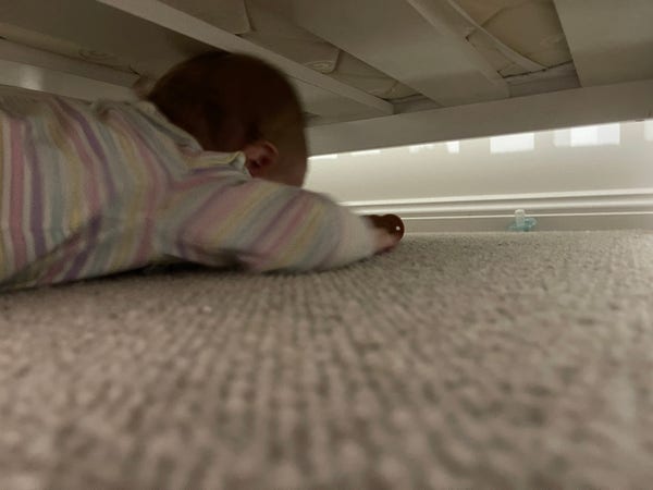 Babby, in a striped long sleeve shirt, crawling on the floor under her cot towards a dropped dummy. 