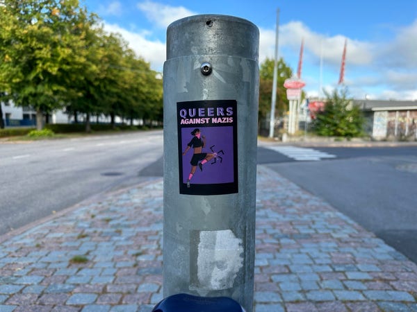 A sticker on a post. Sticker says ”queers against nazis” and has a drawing of a person kicking a swastika