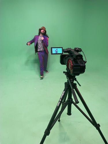 Musical artist Dizraeli on a green screen film set, with a camera in front of him. He is wearing an iridescent purple suit.