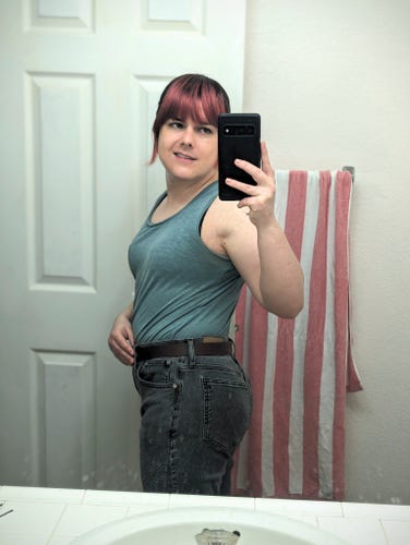 A white trans girl with dyed red hair in a tank top, tucked into fitted jeans and a belt. She's smiling while looking at her phone and twisted to the side to get a look at her butt.