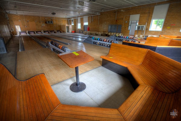 A relatively tidy small bowling alley with six lanes. Benches are in the foreground, and the lanes stretch into the background. 
