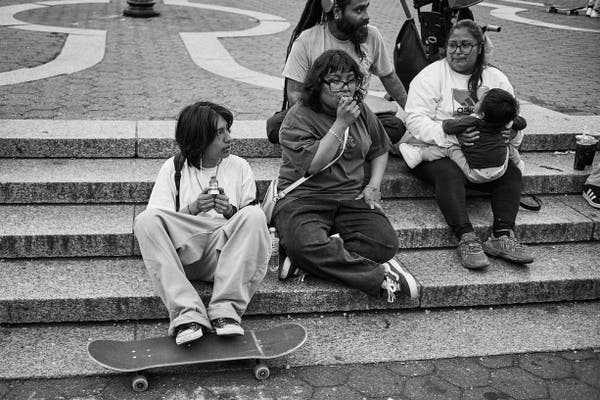 The black and white image depicts four individuals sitting on a staircase, seemingly in an outdoor public space. From left to right: a person perched on a skateboard, with one leg on the board and the other hanging down the step, holding a beverage. Next to them, a person is seated eating, with their gaze down. Behind, an individual with headphones is observing the surroundings. On the far right, another person is holding what appears to be a mobile device close to their face. The atmosphere seems casual, with the individuals engrossed in their own activities.