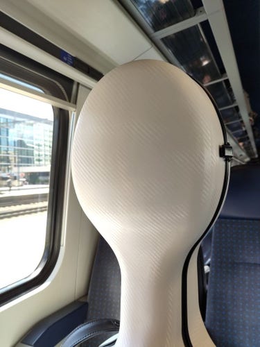Foto des Kopfes eines weissen Cellokastens in einem Zugsabteil mit blauen Sitzen. Durch das Waggonfenster ist ein Ausschnitt eines sonnigen Bahnhofs zu sehen.