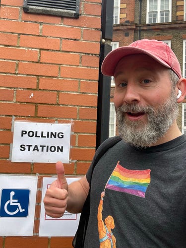 Martin is giving a thumbs-up next to a "POLLING STATION" sign on a brick wall. He wears a cap and a shirt with a rainbow flag graphic.