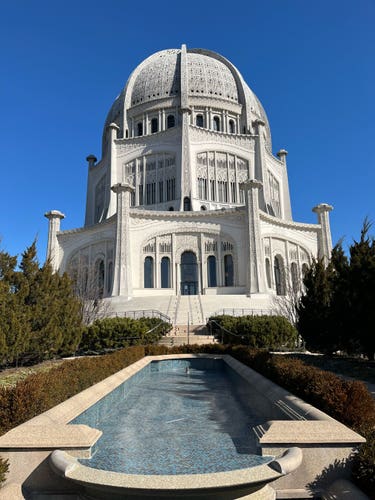 An ornate white building with a large dome and intricate detailing under a blue sky, with a reflecting pool in the foreground.