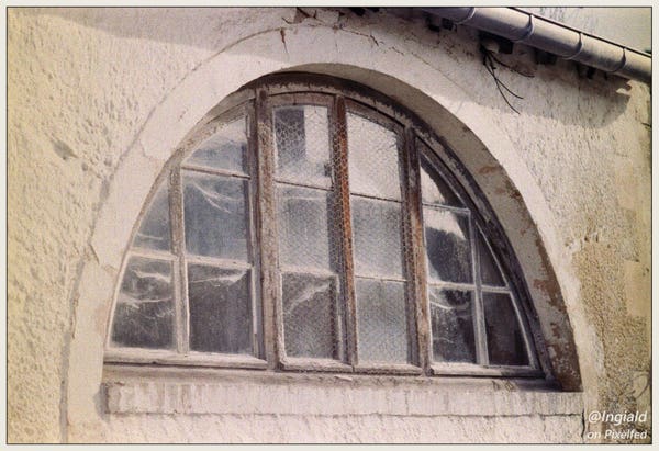 Colour picture of an outside window in a white stucco wall. The window is in the shape of a half circle with the flat side facing down. The window is divided into sixteen smaller panes of glass by a simple rows and columns pattern. The middle six panes are covered by chicken wire. Behind the glass you can see wisps of cobweb. The picture is more grainy than it should be with yellowing to the sides. Possibly indicating it is overexposed and that the camera needs some CLA.