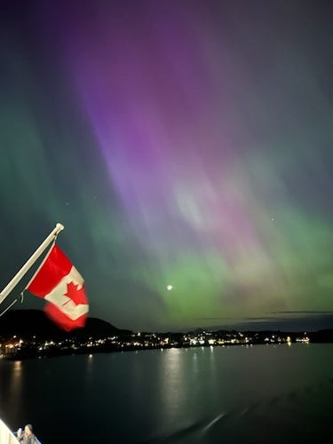 Green and purple lights in the sky, seen over the lights of a small coast city (Bella Coola, I think) from aboard a ferry passing by. A Canadian flag, hanging on the ferry, is in the vorner of the pic
